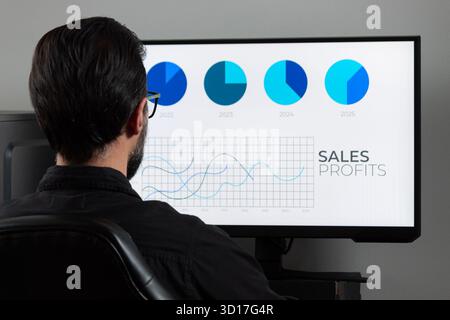 Nähern Sie sich einem Mann hinten links im Bild, der einen Computerbildschirm mit einer blauen Gewinngrafik auf einem pc-Bildschirm ansieht Stockfoto