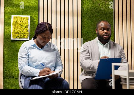 afroamerikanische Bewerber, die im Flur vor dem HR-Büro sitzen und ihre Lebensläufe mit starken Qualifikationen vorbereiten. Rivalen bei der Besetzung der neuen leeren Geschäftsstelle. Stockfoto