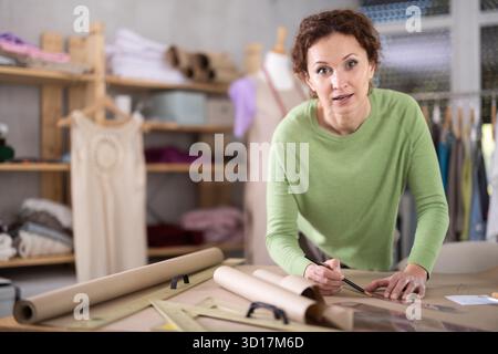 Die Schneiderin zeichnet in der Nähwerkstatt anhand von Mustern eine Skizze der zukünftigen Kleidung auf Papier Stockfoto
