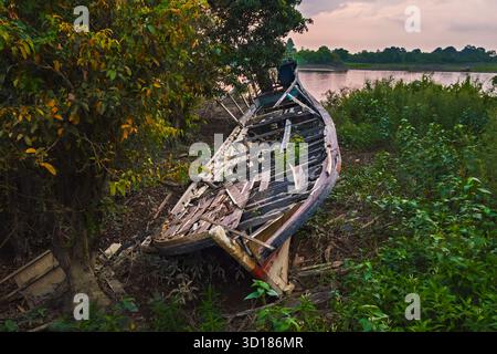 Ein altes Brokenwood-Boot wurde verlassen an einem Flussufer, umgeben von grüner Vegetation und Bäumen. Stockfoto
