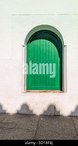 Leuchtend gewölbtes grünes Holzfenster in einer strukturierten weißen Succo-Wand mit starken Schatten auf dem Bürgersteig. Stockfoto