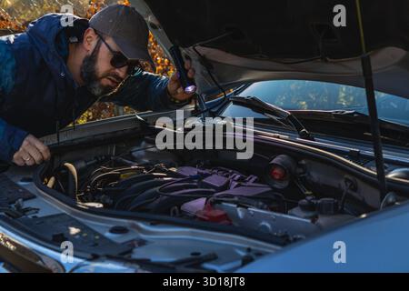 Ein Mann überprüft den Motor seines Autos in sonniger Umgebung. Er konzentriert sich, während er eine Taschenlampe verwendet, um die Komponenten unter der Motorhaube zu betrachten. Stockfoto