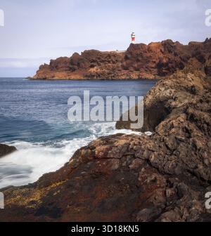 Wellen brechen gegen dunkle Felsen, während der Leuchtturm Punta de Teno in Teneriffa, Spanien, hoch über dem Meereshimmel steht Stockfoto