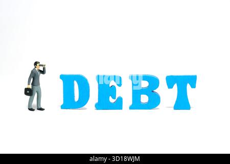 Miniaturmenschen Spielzeug-Fotografie. Ein Geschäftsmann, der ein Fernglas benutzt, steht neben dem Wortbrief aus SCHULDENHOLZ. Isoliert auf weißem Hintergrund. Stockfoto