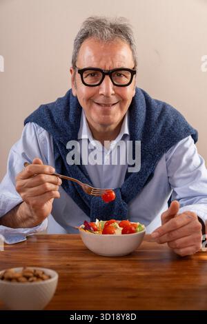 Ein fröhlicher Mann sitzt an einem Holztisch und genießt einen bunten Salat. Er trägt einen blauen Pullover über den Schultern und lächelt warm und sorgt für eine Freude Stockfoto
