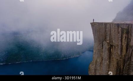 Majestätische Preikestolen Cliff steht hoch über dem Fjord, umgeben von Nebel. Eine einsame Figur genießt die spektakuläre Aussicht, die ein Gefühl von Abenteuer und Ruhe in der atemberaubenden Landschaft Norways weckt. Stockfoto