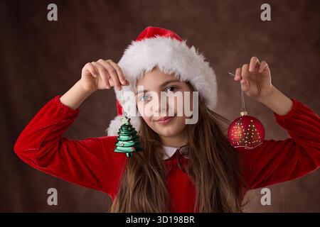 Mädchen halten in Händen Weihnachtsbaumkugeln Dekoration und gekleidet in Santa Claus Hut, Weihnachtsporträt auf rotem Hintergrund Stockfoto