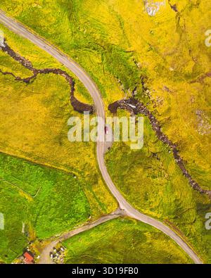 Die Aussicht auf ein einsames rotes Auto aus der Vogelperspektive liegt an der Kreuzung der gewundenen Straßen durch die lebhaften gelben und grünen Hügel, Norðradalur, Streymoy, Färöer Inseln. Stockfoto