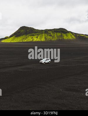 Aus der Vogelperspektive eines einsamen Autos durchquert die krass schwarze vulkanische Landschaft, die vom lebhaften grünen Vulkan Maelifell in Skaftárhreppur, Island, im Schatten steht. Stockfoto
