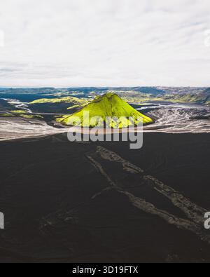 Aus der Vogelperspektive erhebt sich der moosbedeckte Vulkankegel von Mælifell dramatisch aus dem harten, schwarzen Sand, einem lebendigen grünen Leuchtfeuer vor der einsamen Landschaft, Maelifell, Skaftárhreppur, Island. Stockfoto