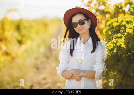 Frau in Hut und Sonnenbrille, die auf dem Feld des üppigen Laubs von Trauben steht Stockfoto