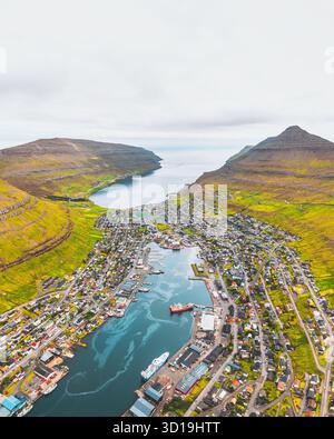 Blick aus der Vogelperspektive auf die Stadt eingebettet zwischen grünen Hügeln und dem blauen Fjord, den Hafen voller Schiffe unter bewölktem Himmel, Klaksvík, Nördliche Inseln, Färöer Inseln. Stockfoto
