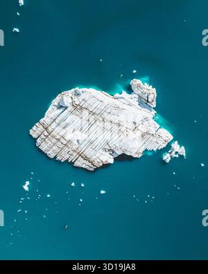 Aus der Vogelperspektive auf einen einsamen Eisberg, der im türkisfarbenen Wasser liegt, dessen strukturierte Oberfläche unter der Sonne glänzt, Jökulsárlón, Sveitarfélagið Hornafjörður, Island. Stockfoto