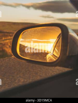 Blick auf einen goldenen Sonnenuntergang, der im Seitenspiegel eines Autos reflektiert und ein warmes Leuchten über die Asphaltstraße und die entfernten grünen Felder in Ingjaldshólskirkja, Snæfellsbær, Island ausstrahlt. Stockfoto