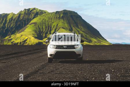 Maelifell, Island - 28. August 2025: Der Blick auf einen scharf weißen SUV steht im Kontrast zur dunklen vulkanischen Erde, einem grünen Berg, der sich im Hintergrund erhebt. Stockfoto