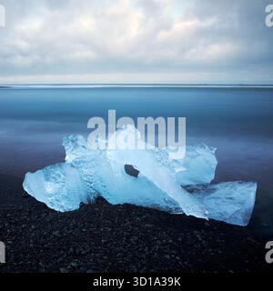 Blick auf durchscheinendes blaues Eis, das auf dem schwarzen Sand vor dem Hintergrund eines stimmungsvollen Ozeans und Himmels liegt, Jokulsarlon, Sveitarfélagið Hornafjörður, Island. Stockfoto