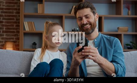 Glückliche Familie im Wohnzimmer Vater mit niedlichem Mädchen Kind, das Telefon sieht, kommuniziert über mobile Social-Media-Inhalte, entspannt auf der Couch zu Hause Stockfoto