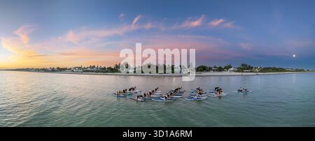 Aus der Vogelperspektive sehen Sie eine Gruppe von Menschen, die auf Paddelbrettern im türkisfarbenen Meer Yoga machen, vor dem Hintergrund weißer Strände und lebendiger Sonnenuntergangshimmel, Progreso, Yucatan, Mexiko. Stockfoto
