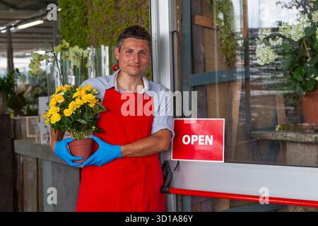 Ein Mann mit Schürze und Handschuhen, der eine gelbe Begonienpflanze hält und an einer offenen Ladentür steht Stockfoto