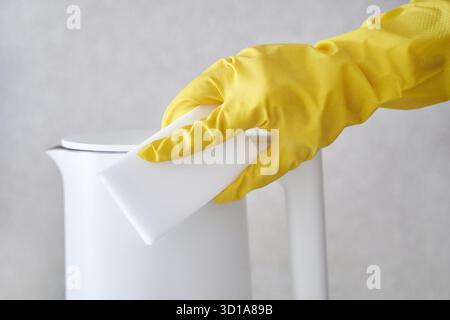 Person reinigt weißen Wasserkocher mit gelbem Handschuh und Schwamm für Küchenpflege und Hygiene. Stockfoto