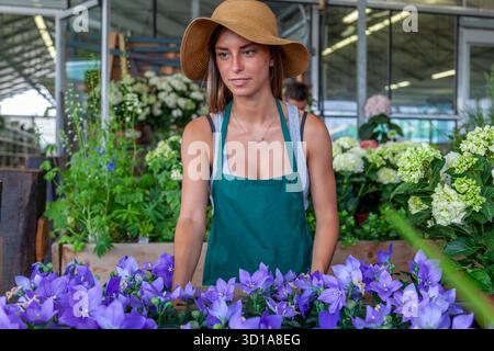 Frau in Schürze und Strohhut, die zwischen verschiedenen Pflanzen und Blumen in einem Gartencenter steht Stockfoto