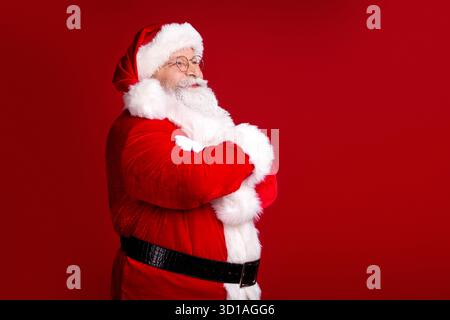 Der Weihnachtsmann steht selbstbewusst mit überkreuzten Armen in einem roten Anzug und weißem Pelz vor einem auffälligen roten Hintergrund für festliches Weihnachtsmarketing Stockfoto