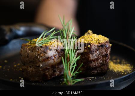Nahaufnahme von zwei gegrillten Rindfleischfilet Mignon-Steaks garniert mit Rosmarin und Gewürzen, serviert auf einem schwarzen Teller. Stockfoto