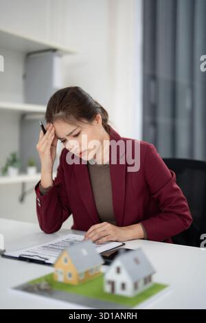Asiatische Agentin hat Stress beim Überprüfen der Papiere für Immobilienverträge mit Hausmodellen am Schreibtisch professionelle Geschäftsentscheidungen stellen die Büroarbeit in Frage Stockfoto