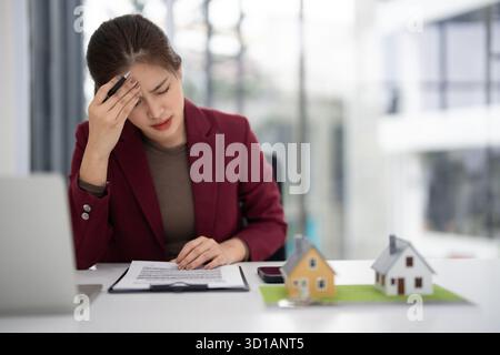 Gestresste Erwachsene asiatische Immobilienmakler, die Immobiliengeschäfte planen. Sie fühlt sich von Hypothekendokumenten oder einer Strategie für den Verkauf von Häusern überwältigt. Stockfoto