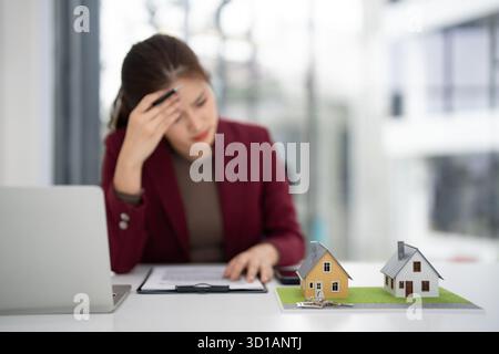Asiatische Erwachsene Immobilienexperten fühlen sich überfordert, Immobilienmarktprobleme in einer modernen Büroumgebung mit Hausmodellen zu betrachten. Stockfoto