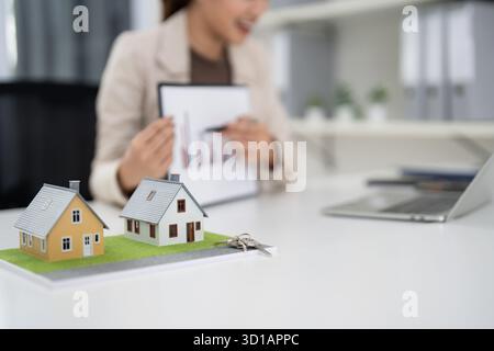 "Professioneller asiatischer Immobilienmakler für Erwachsene erklärt Finanzdiagramme für den Immobilienerwerb. Miniaturhäuser und Schlüssel werden zur Prospektion auf dem Schreibtisch angezeigt Stockfoto