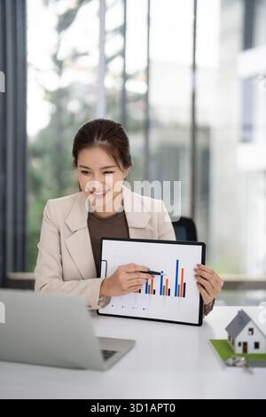 Erwachsene asiatische Frau erklärt potenziellen Käufern zuversichtlich Immobilienmarktanalysen Immobilienmakler diskutiert Investitionswert in einem hellen Büro Stockfoto