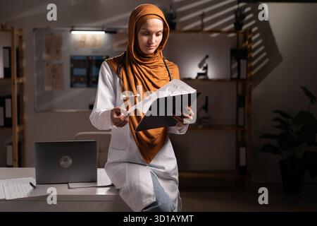 Selbstbewusste muslimische Ärztin, die Hijab trägt und Patienteninformationen auf dem Klemmbrett überprüft, die in der Nachtschicht im Krankenhaus oder in der Klinik arbeitet. Medizin, hea Stockfoto