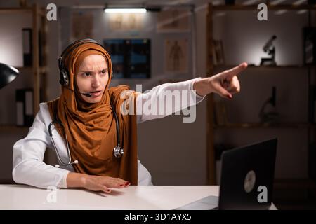 Wütende muslimische Ärztin, die Hijab und Stethoskop trägt, schreiend und auf dich gerichtet, während sie an der Nachtschicht im Krankenhaus arbeitet. Gesundheitsmedizin c Stockfoto