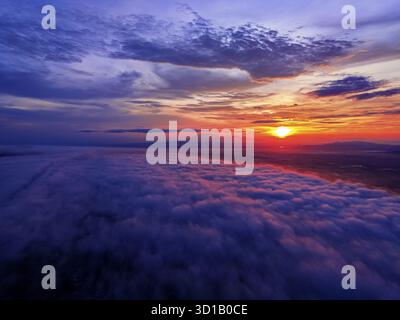 Aus der Vogelperspektive auf einen atemberaubenden Sonnenaufgang über einer dicken Wolkendecke, mit dem strahlenden Licht der Sonne im Kontrast zur fernen Silhouette des Lawu Mountain, Karanganyar, Zentral-Java, Indonesien. Stockfoto