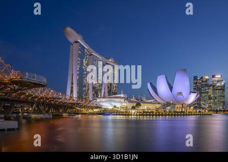 Blick auf die beleuchtete Helix Bridge, die sich im Wasser spiegelt und zum berühmten Marina Bay Sands und ArtScience Museum unter einem dunkelblauen Himmel führt, Singap Stockfoto