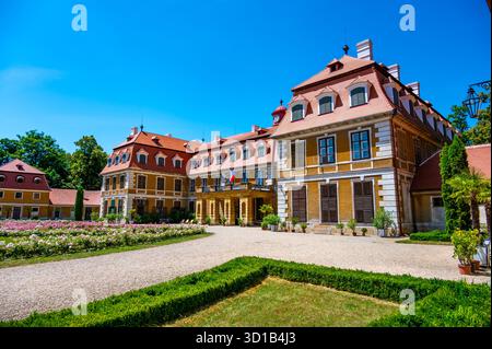 Panoramablick auf das Schloss Rajec Jestrebi an einem sonnigen Sommertag mit blauem Himmel, blühenden rosa Rosen und einem Brunnen vor dem historischen Gebäude. Stockfoto