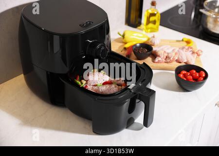 Air Fritteuse mit rohen Hähnchenstangen, Schenkeln, Gemüse und Gewürzen auf weißer Marmorplatte, Nahaufnahme Stockfoto