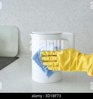 Person, die den weißen Wasserkocher mit gelben Handschuhen und blauem Tuch auf der Küchenzeile reinigt. Stockfoto