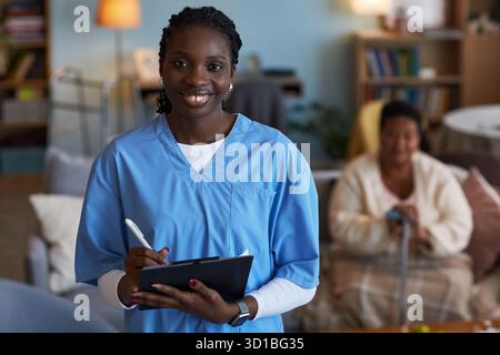 Porträt der jungen erwachsenen Schwarzen Frau, die medizinische Peelings trägt, lächelnd in die Kamera, die Klemmbrett und Stift hält, ältere Schwarze Frau, die im Hintergrund mit Gehstock im Wohnzimmer sitzt, während des Patientenbesuchs Stockfoto