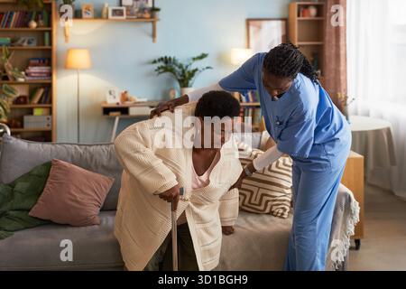 Ältere Schwarze Frau, die Stock benutzt und Hilfe von einer jungen erwachsenen Schwarzen Krankenschwester in Peelings erhält, die ihr beim Aufstehen vom Sofa im Wohnzimmer hilft, während des Hausbesuchs Stockfoto