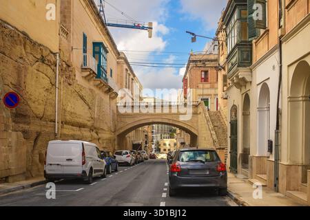 Enge maltesische Straße mit Kalksteinbauten, bunten Holzbalkonen, Steintreppen, geparkten Autos, Straßenschildern, und eine Bogenbrücke Stockfoto