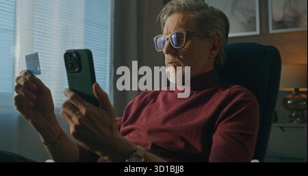 Alter Mann in der Brille verwendet Smartphone und Kreditkarte vollständige Online-Transaktion, während Sie sich im Hotelstuhl entspannen. Concept Secure Online Shopping, Komfortable Digitale Zahlungen, E-Commerce Auf Reisen. Stockfoto