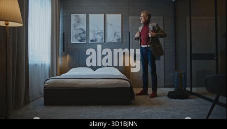 Ein Schöner Reifer Mann Checkt In Ein Modernes Hotelzimmer Ein. Geschäftsmann zieht die Jacke aus und legt sich erschöpft auf das Bett. Konzept für Geschäftsreise, Hotelaufenthalt und Erholung nach der Reise. Statische Aufnahme. Stockfoto