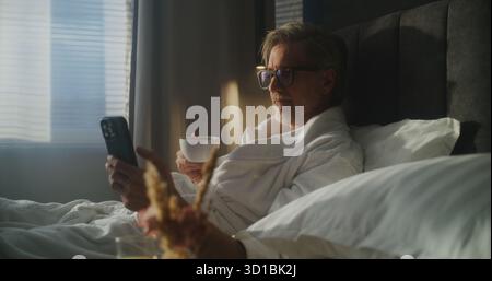 Ein reifer Executive sitzt im Hotelbett, trägt Bademantel und überprüft das Smartphone, während er eine Tasse Kaffee zum Frühstück hält. Work Life Integration, Mobile Technologie Verwenden, Geschäftsreise. Dolly Shot. Stockfoto