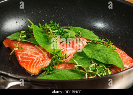 Erhöhte Sicht auf zwei Lachsfilets, die mit frischem Salbei und Thymian in einer schwarz beschichteten Pfanne mariniert sind. Stockfoto
