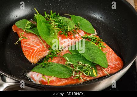 Erhöhte Sicht auf zwei Lachsfilets, die mit frischem Salbei und Thymian in einer schwarz beschichteten Pfanne mariniert sind. Stockfoto