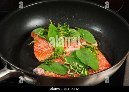 Erhöhte Sicht auf zwei Lachsfilets, die mit frischem Salbei und Thymian in einer schwarz beschichteten Pfanne mariniert sind. Stockfoto