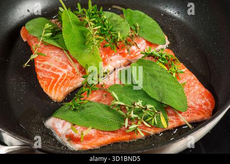 Erhöhte Sicht auf zwei Lachsfilets, die mit frischem Salbei und Thymian in einer schwarz beschichteten Pfanne mariniert sind. Stockfoto