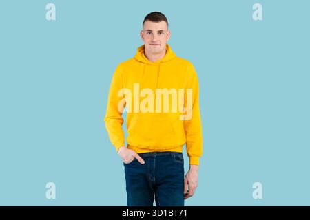 Junger Mann in Yellow Hoodie posiert selbstbewusst vor hellblauem Hintergrund in einer legeren Umgebung Stockfoto